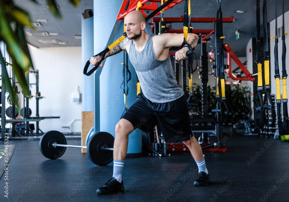 Φωτογραφία Tattooed athletic man exercising with suspension fitness rope in the gym