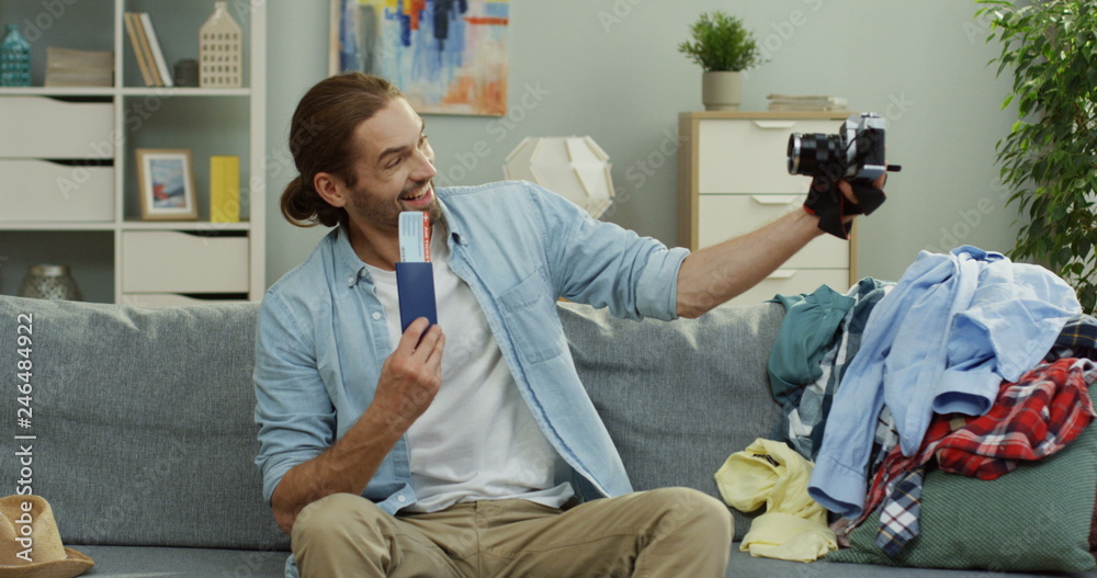 Obraz premium Attractive and cheerful young Caucasian man taking selfie photos on the camera of him holding a passport with a ticket while sitting on the sofa among mess before traveling.