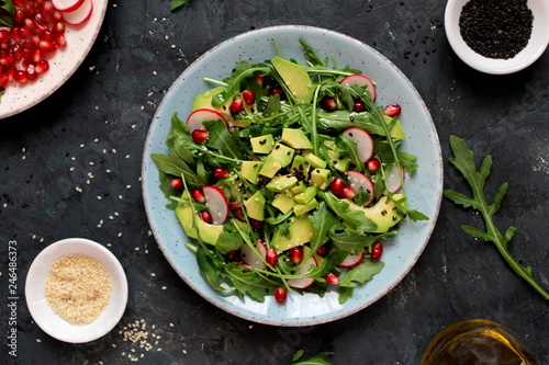Ingredient for making salad on dark background. Vegetable salad in plate, avocado, arugula, pomegranate, radish and sesame. Top view with copy space. Vegetarian and vegan food.