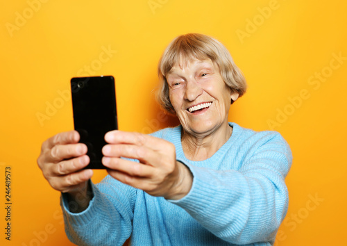 lifestyle, tehnology and people concept: grandma in blue sweater smiles and takes a selfie over yellow background