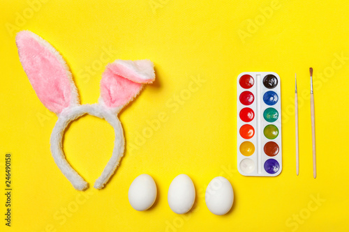 Happy Easter concept. Preparation for holiday. Decorative eggs colorful paints and bunny ears furry costume toy isolated on trendy yellow background. Simple minimalism flat lay top view copy space