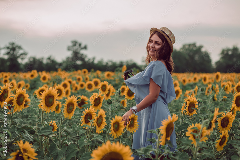 Dreaming young woman in blue dress and hat holding a cup of coffee in a field of sunflowers at summer, view from her side. Looking to the side. Smiling Dreaming young woman in blue dress and hat holding a cup of coffee in a field of sunflowers at summer, view from her side. Looking to the side. Smiling