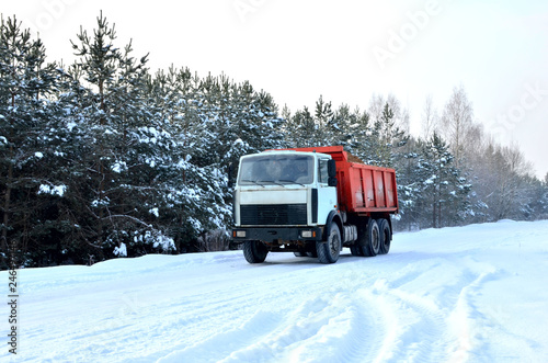 Cargo dump truck rides on a snowy road to load sand into career. Winter road against the backdrop of fabulous spruce trees in the snow and blue sky