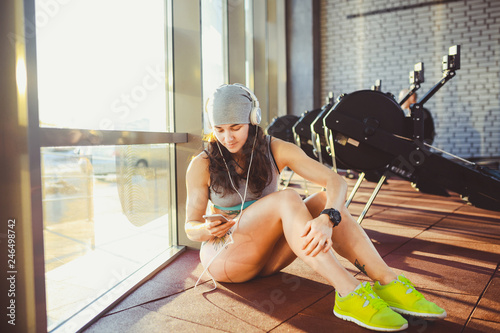 Theme sports, health and technology. beautiful sexy Caucasian woman sportswoman in gray sportswear and hat sits by window with sun setting uses smartphone listen music in large headphones on head