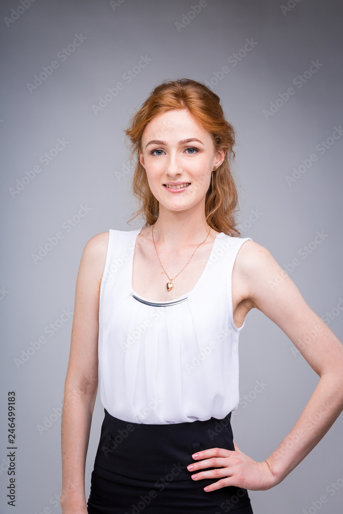 portrait vertical up to the waist young, beautiful business woman, student with lred, curly hair and freckles on gray background. SHe dressed in a white blouse with short sleeves about open shoulders portrait vertical up to the waist young, beautiful business woman, student with lred, curly hair and freckles on gray background. SHe dressed in a white blouse with short sleeves about open shoulders