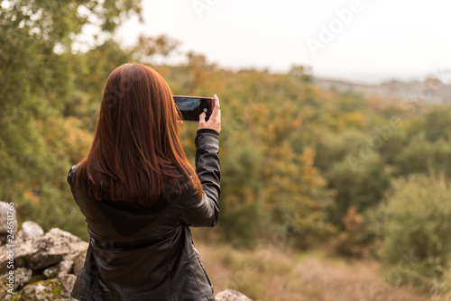 A young red-haired woman takes pictures with her mobile phone in a forest