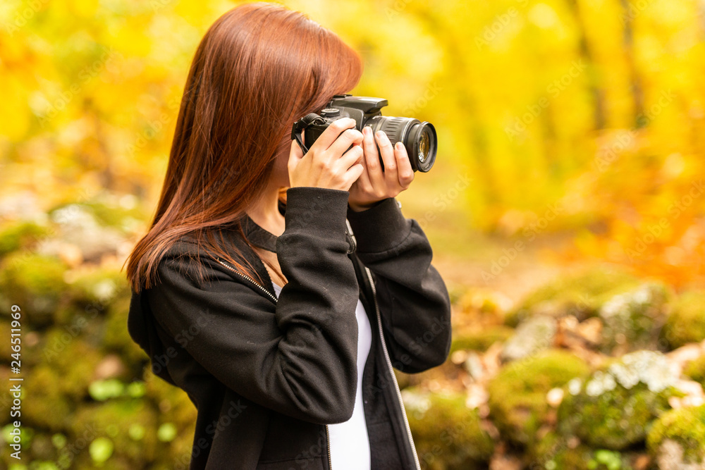 A young woman with reddish hair takes pictures with a reflex camera in a forest 