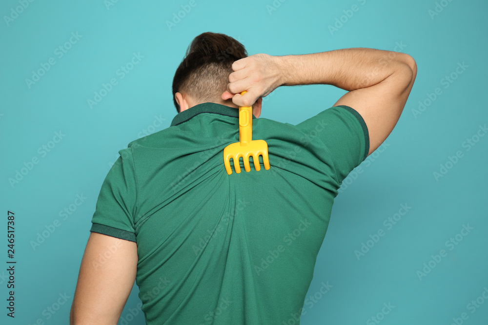 Young man scratching back with toy rake on color background. Annoying itch - obrazy, fototapety, plakaty 
