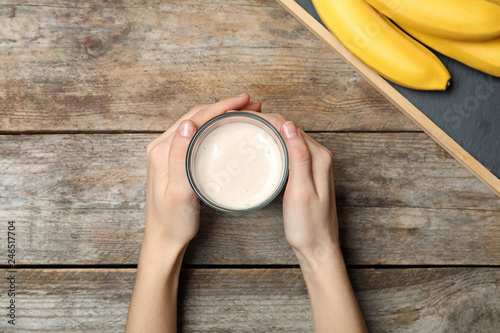 Woman holding glass of healthy protein shake near bananas at wooden table, top view