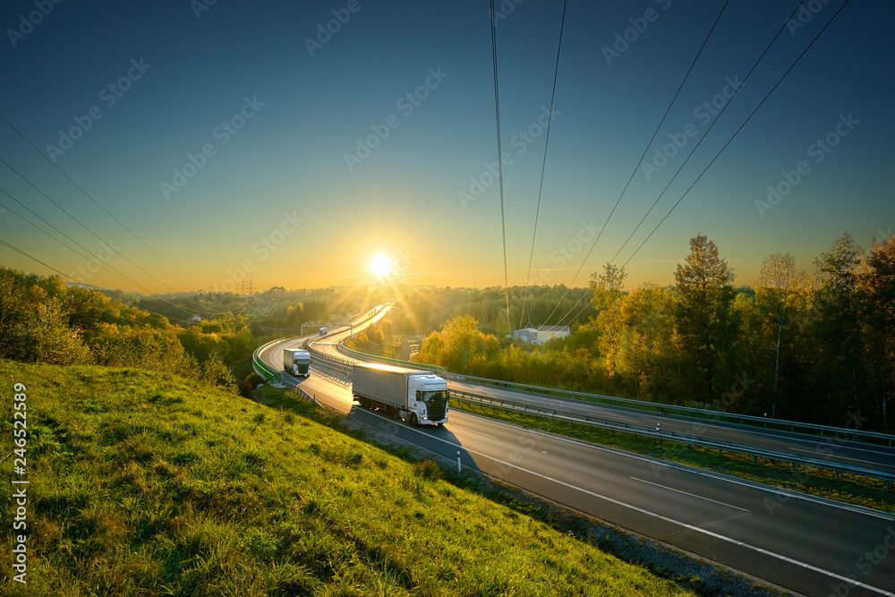 Three white trucks driving on the asphalt highway in autumn landscape in the rays of the sunset