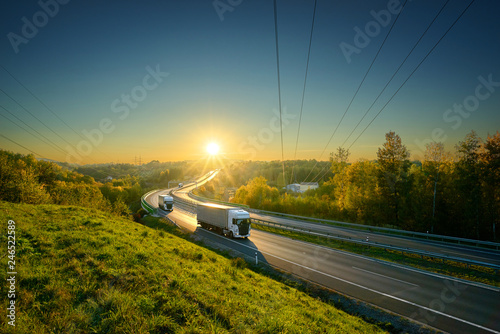 Three white trucks driving on the asphalt highway in autumn landscape in the rays of the sunset