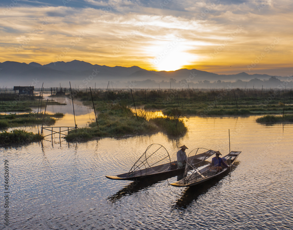 Obraz premium two Intha fishermen warming themselves by the fire on their boats during early morning sunrise on Lake Inle, Myanmar, Burma