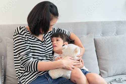 Asian mother embrace little preschool frustrated kid sitting on couch together at home.