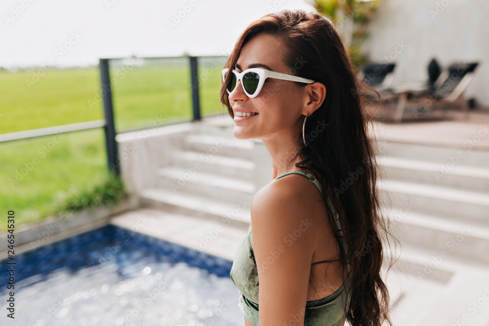 Close-up portrait of attractive happy smiling woman in glasses and swimsuit posing during photoshoot on background of green field and pool. Girl relaxing on spa resort in good sunny day Close-up portrait of attractive happy smiling woman in glasses and swimsuit posing during photoshoot on background of green field and pool. Girl relaxing on spa resort in good sunny day