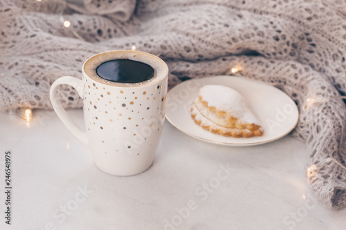 Warm cozy knitwear and a cup of coffee with a cake on white marble windowsill against white window background. Copy space.