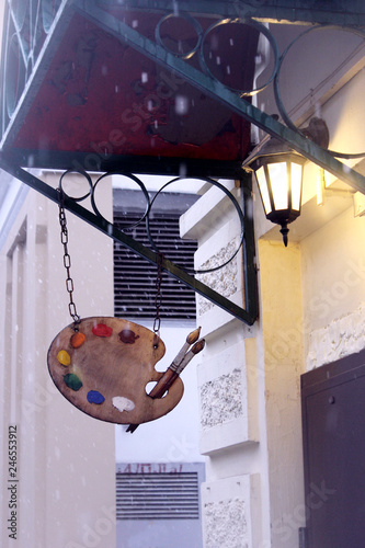 Hanging sign of an art shop of a wooden palette with bright colors and tassels: winter evening, lantern light and falling  snow