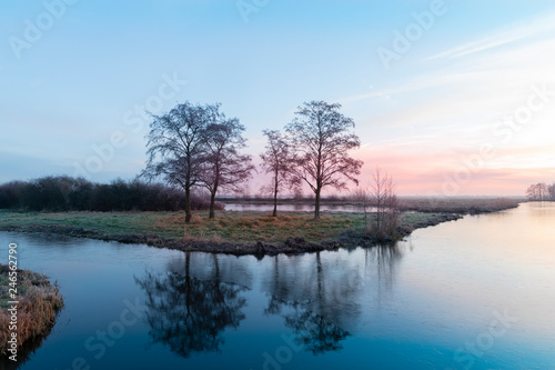 Piece of land in a semi-frozen ditch with bare tree silhouettes in winter morning during sunrise.