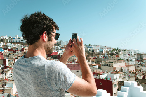 Portrait of happy young man on Morocco, tourist with smartphone taking pictures of old city Tangier from terrace
