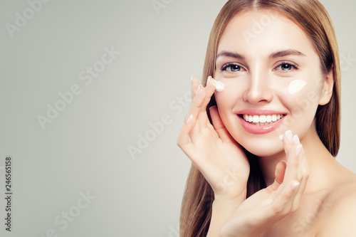 Exited beautiful woman with healthy clear skin applying moisturizing cream on her face, closeup portrait. Skin care, beauty and facial treatment concept
