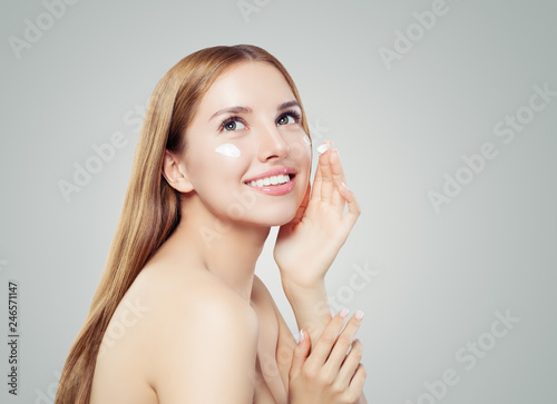 Cheerful woman applying moisturizing cream on her face. Happy model girl with healthy clear skin on white background