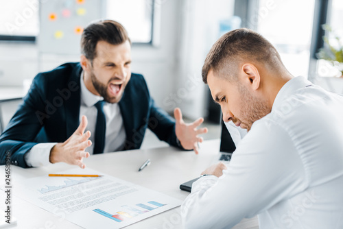 selective focus of upset man sitting near angry man in formal wear screaming in modern office