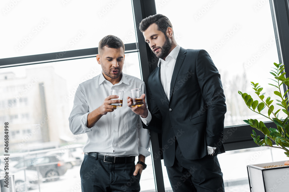 businessmen standing with hands in pockets while holding glasses with alcohol drinks