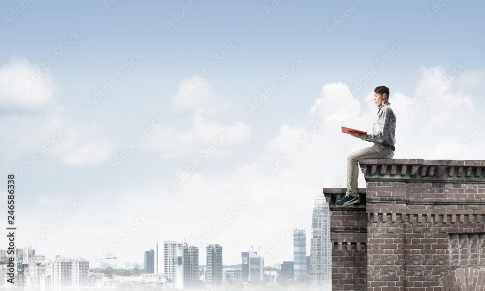 Man on roof edge reading book and cityscape at background