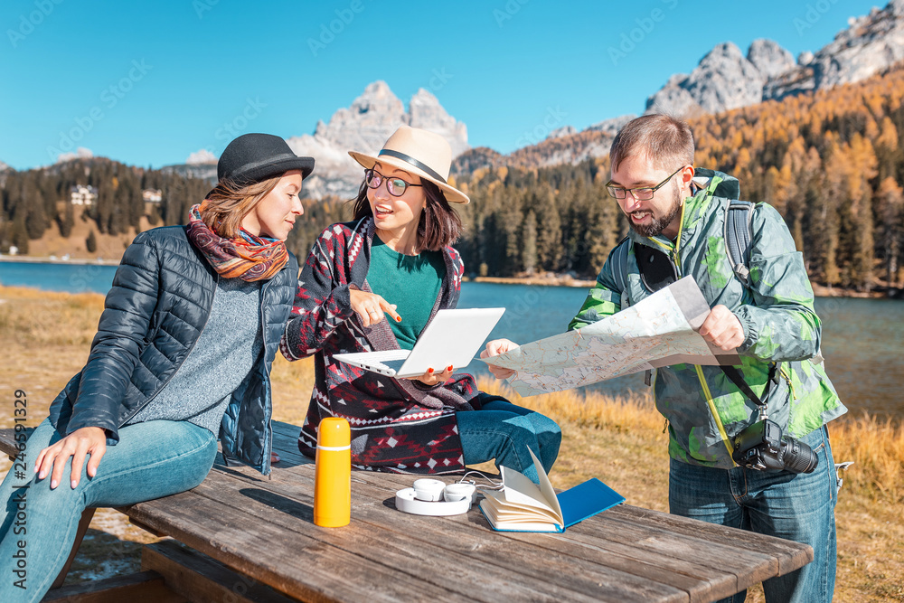 A group of friends with a map and a laptop in a mountain reserve on the shore of a beautiful lake are looking for a good hotel to book on the Internet to continue the adventure A group of friends with a map and a laptop in a mountain reserve on the shore of a beautiful lake are looking for a good hotel to book on the Internet to continue the adventure