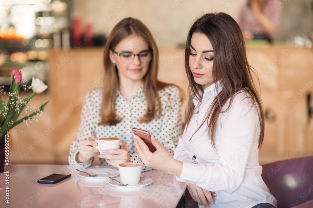 Two girls sitting in a cafe drinking cappuccino and using smartphone. Modern girl