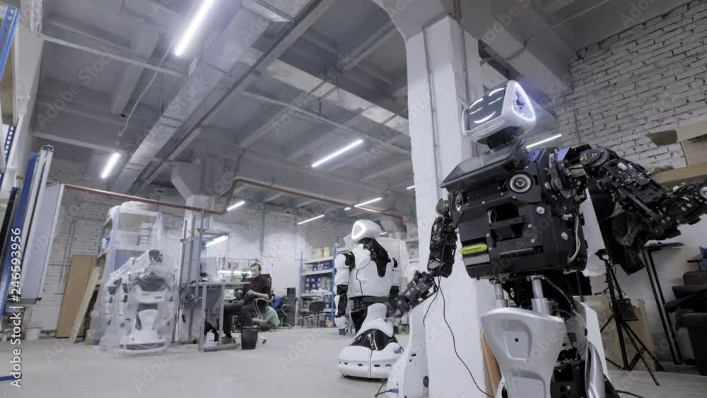 Factory for the production of robots, modern robotic developments. The disassembled robot waves his hands in greeting and dances in the workshop. Engineers walk in the background Factory for the production of robots, modern robotic developments. The disassembled robot waves his hands in greeting and dances in the workshop. Engineers walk in the background