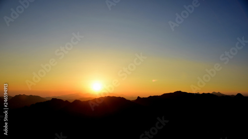 Picture of a sun setting behind a dense forest area followed by mountains.