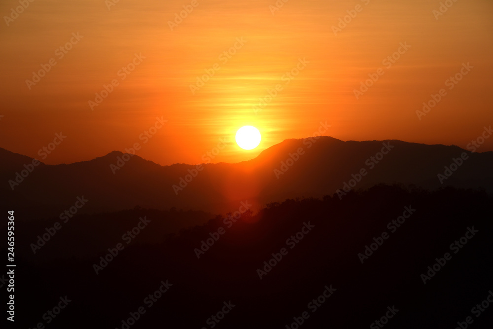 Picture of a sun setting behind a dense forest area followed by mountains.