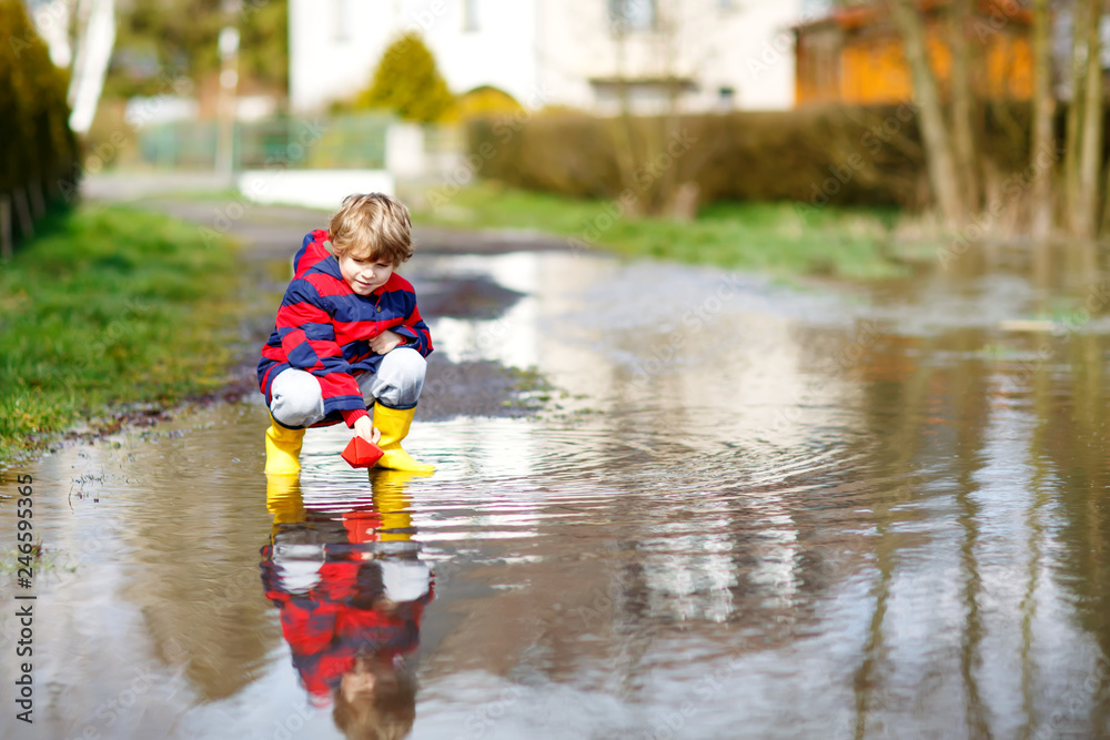 Happy little kid boy in yellow rain boots playing with paper ship boat by  huge puddle