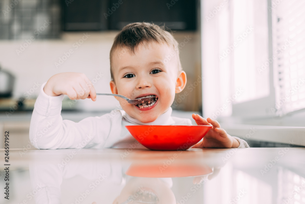 A child in the kitchen eating their own oatmeal with a red plate A child in the kitchen eating their own oatmeal with a red plate