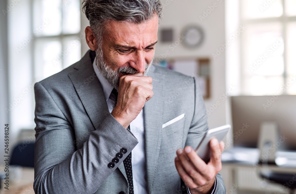 A frustrated businessman with smartphone standing in an office, reading bad news. A frustrated businessman with smartphone standing in an office, reading bad news.
