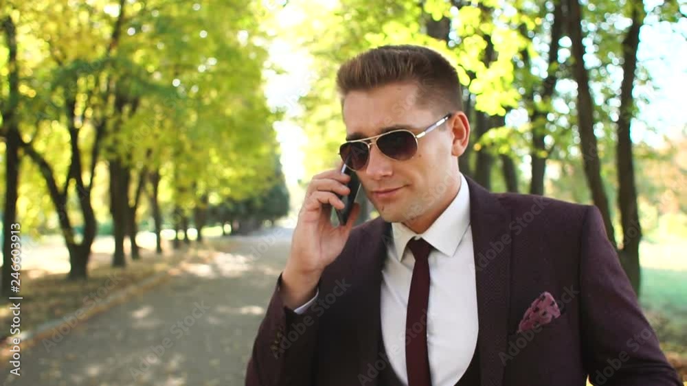 Handsome young stylish male businessman talking on the phone on a walk in the autumn park on the background of yellow leaves, dressed in a stylish business suit and sunglasses. Handsome young stylish male businessman talking on the phone on a walk in the autumn park on the background of yellow leaves, dressed in a stylish business suit and sunglasses.