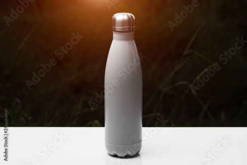 Stainless thermo bottle for water, tea and coffe, on white table. Dark grass background with sunlight effect. Thermos silver color. © Lalandrew