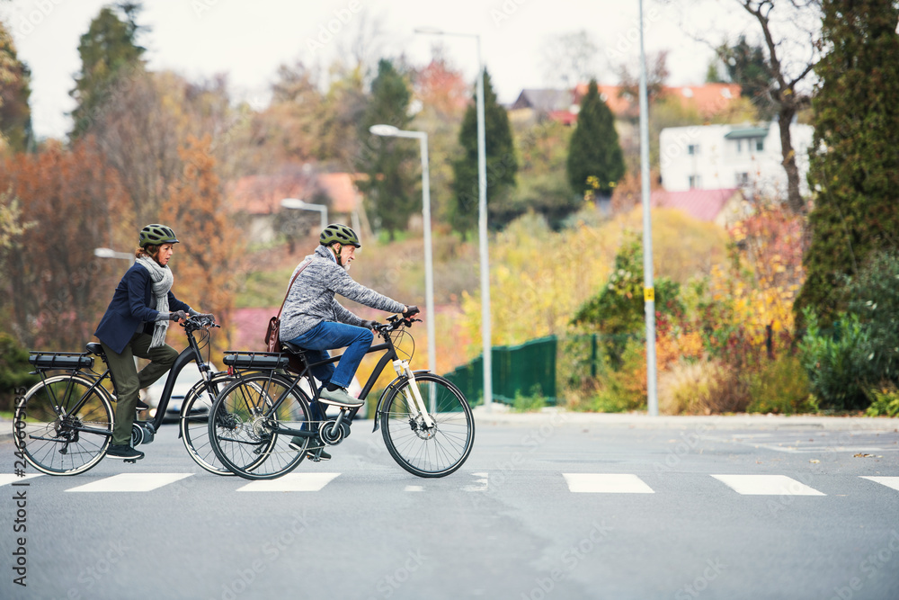 Obraz premium Active senior couple with electrobikes outdoors crossing a road in town.