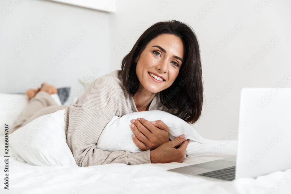 Photo of smiling woman 30s using laptop, while lying in bed with white linen in bright room Photo of smiling woman 30s using laptop, while lying in bed with white linen in bright room