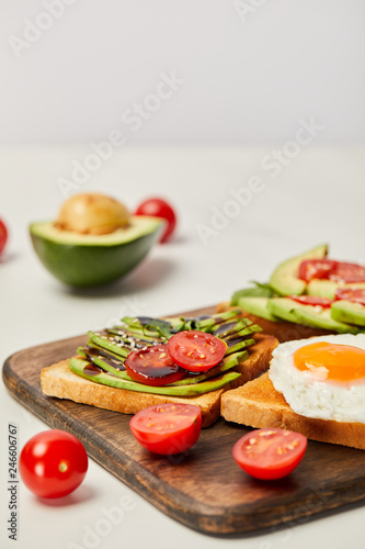selective focus of wooden cutting board with toasts, scrambled egg, cherry tomatoes and avocado on grey background
