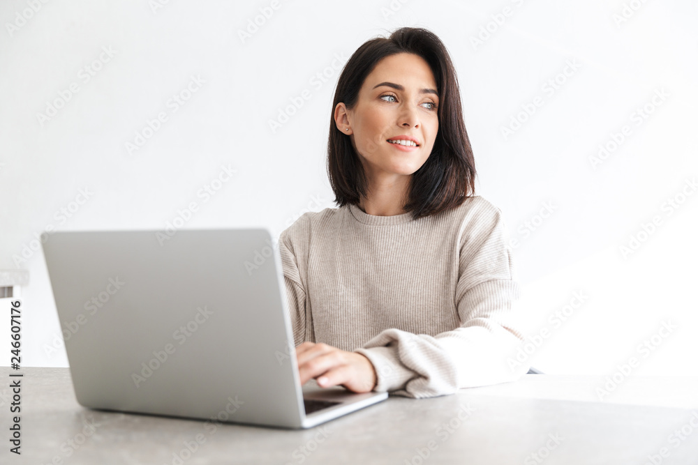 Fototapeta premium Image of beautiful woman 30s working on laptop, while sitting over white wall in bright room