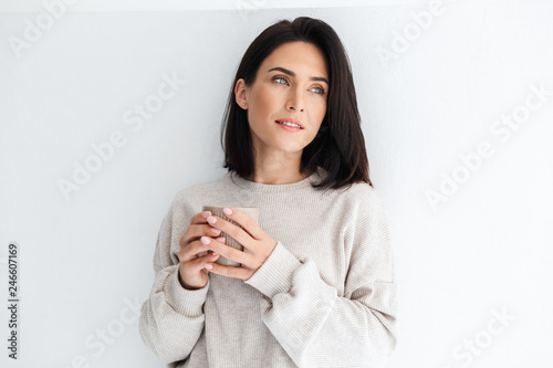 Photo of gorgeous woman 30s holding cup with tea, while standing over white wall indoor