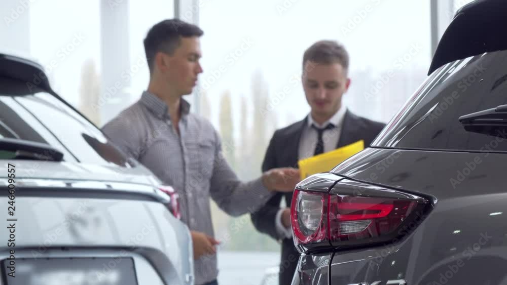 Selective focus on a car, male customer talking to auto dealer on the background. Man buying new automobile, getting an advice from salesman. Ownership, insurance concept Selective focus on a car, male customer talking to auto dealer on the background. Man buying new automobile, getting an advice from salesman. Ownership, insurance concept