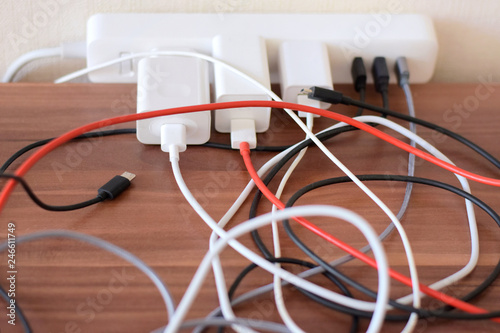 Blurred tangled colorful electricity, usb cables on wooden table. White socket set with mess connection wires for charging gadgets with selective focus. Electrical power outlet 