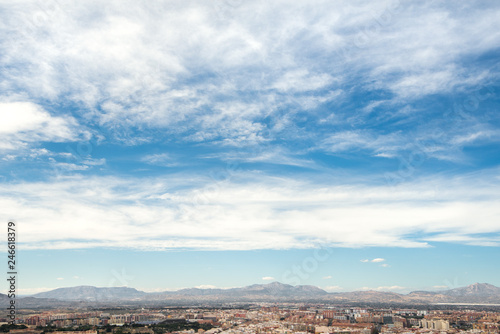 Panoramic view of city from Santa Barbara Castle in Alicante, Spain. Block apartment buildings, parks, roads, houses, palm trees. Beautiful mountain landscape in background, blue sky 