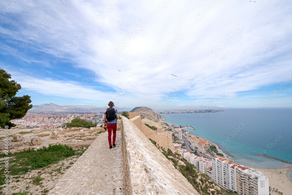 Young man seeing panoramic view of Postiguet beach  from Santa Barbara Castle in Alicante, Spain. Sunny day at Mediterranean sea. Block apartment buildings in a row. Palm trees and vibrant blue water.
