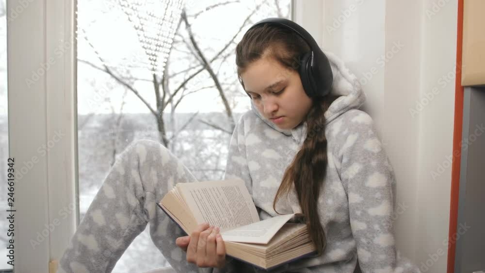 Girl teenager in pajamas and wireless headphones reading a book sitting on the windowsill. Girl teenager in pajamas and wireless headphones reading a book sitting on the windowsill.