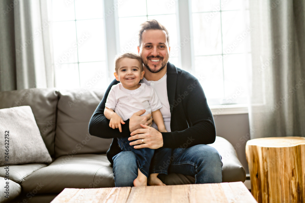 Portrait of handsome father and his cute son hugging, looking at camera and smiling while sitting on sofa at home Portrait of handsome father and his cute son hugging, looking at camera and smiling while sitting on sofa at home