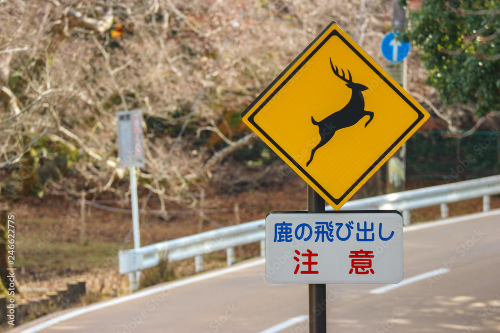 A yellow Japanese language road traffic sign of beware jumping wild deer into the road at nara national park.