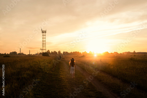 A girl with a floral wreath on her head is walking barefoot across the field at sunset.